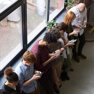 Eine Gruppe Menschen steht aufgereiht vor einem Fenster, alle blicken auf ihr Smartphone