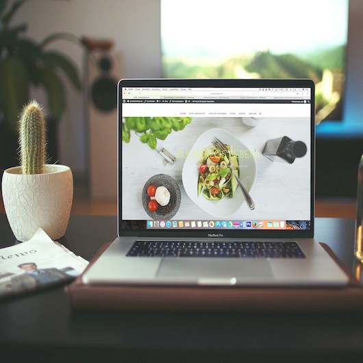 ein Laptop zeigt auf dem Bildschirm Food-Content, daneben ein Kaktus und eine Tageszeitung